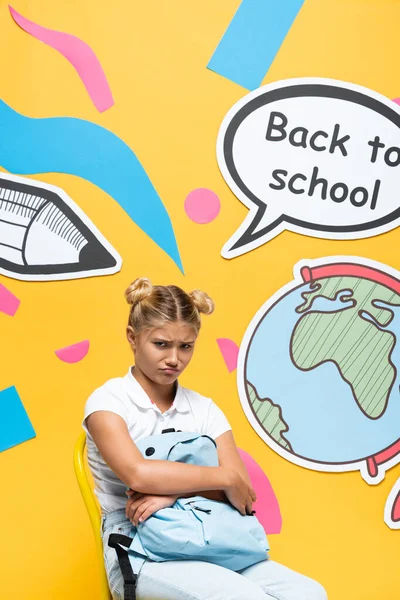 Upset schoolgirl holding backpack on chair near back to school lettering on speech bubble and paper art on yellow background — Stock Photo