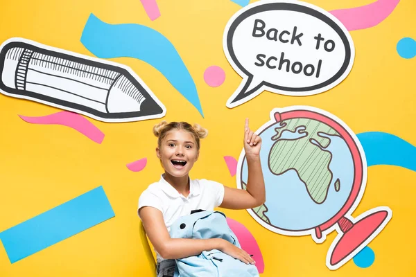 Schulkind mit Rucksack, der auf Schulbuchstaben und Papierelemente auf gelbem Hintergrund zeigt — Stockfoto