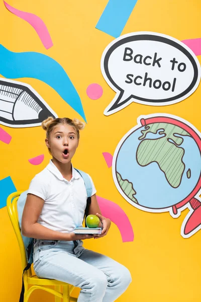 Schockiertes Schulkind hält Buch und Apfel auf Stuhl neben Papierkunst und Sprechblase mit Schulbuchstaben auf gelbem Hintergrund — Stockfoto