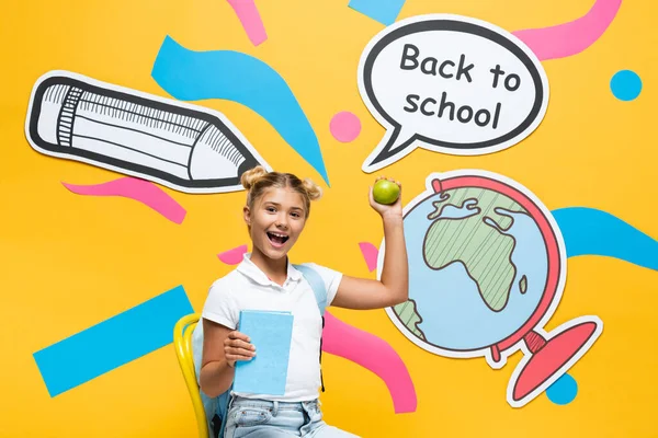 Excited schoolkid with apple and book sitting near back to school lettering on speech bubble and paper art on yellow background — Stock Photo