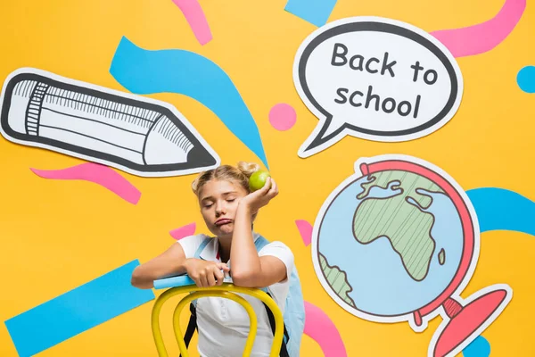 Tired schoolgirl with apple and book sitting near paper elements and speech bubble with back to school lettering on yellow background — Stock Photo