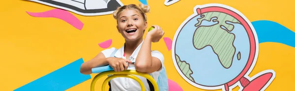 Panoramic crop of excited schoolgirl holding book and apple near paper art on yellow background — Stock Photo