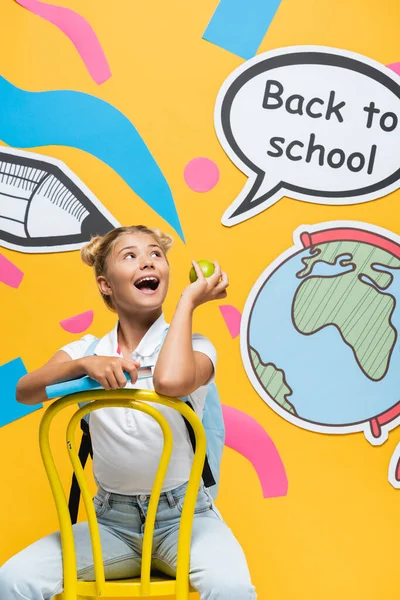 Захоплена дитина з яблуком і книгою, дивлячись на бульбашку мовлення зі спиною до шкільного написання та паперове мистецтво на жовтому тлі — стокове фото