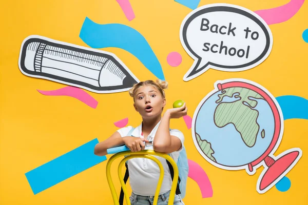 Geschocktes Kind mit Buch und Apfel sitzt neben Sprechblase mit Schulbuchstaben und Papierbasteln auf gelbem Hintergrund — Stockfoto