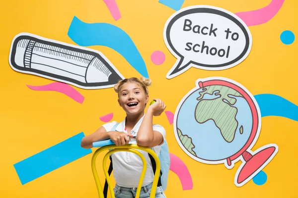 Schoolkid holding book and apple near paper speech bubble and artwork on yellow background — Stock Photo