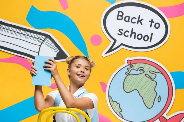 Школьная девочка держит книгу рядом с пузырьком речи со школьными надписями и бумажными рисунками на желтом фоне — стоковое фото