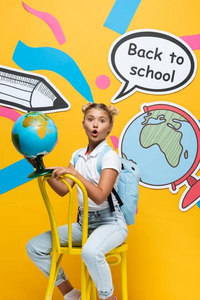 Emocionado colegial sosteniendo globo en silla cerca de elementos de papel sobre fondo amarillo - foto de stock