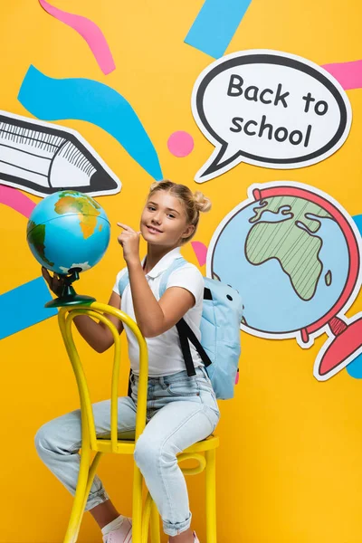 Écolière pointant vers le globe près de l'art du papier avec lettrage de retour à l'école sur fond jaune — Photo de stock