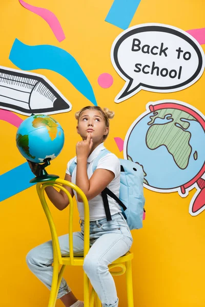 Pensativo niño sosteniendo globo cerca de la burbuja del habla con letras de vuelta a la escuela y el arte del papel sobre fondo amarillo - foto de stock