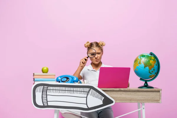 Schoolkid holding loupe near globe, laptop and paper art on desk on pink background — Stock Photo