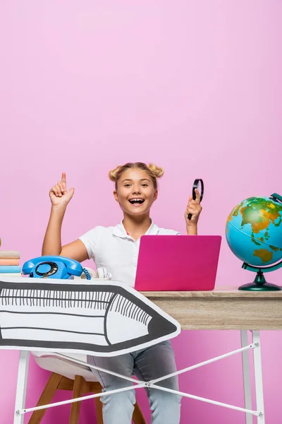 Ребенок показывает пальцем, держа увеличительное стекло возле глобуса, ноутбук и бумажный элемент на розовом фоне — стоковое фото