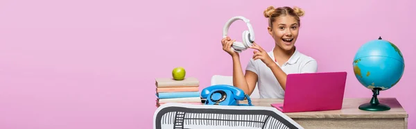 Foto panorâmica de menina segurando fones de ouvido perto do globo, laptop e arte de papel na mesa isolada em rosa — Fotografia de Stock