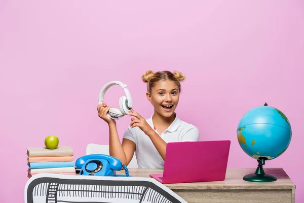 Chica sosteniendo auriculares cerca de la computadora portátil, libros y arte de papel en el escritorio aislado en rosa - foto de stock