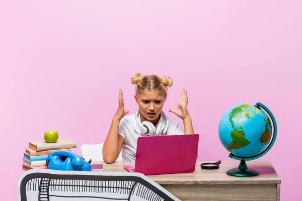 Chica enojada en los auriculares mirando portátil cerca de libros y arte de papel en el escritorio aislado en rosa - foto de stock