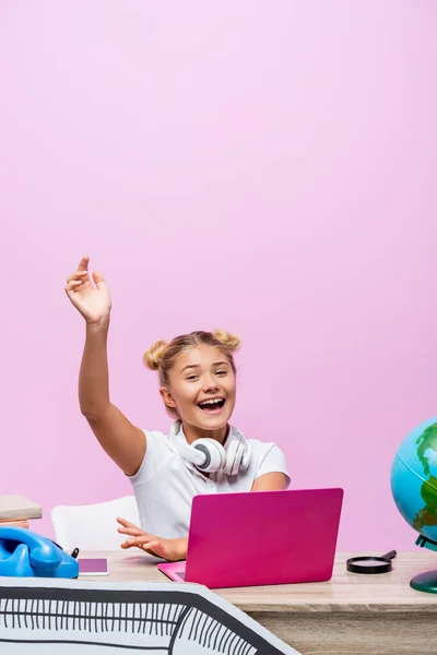 Colegiala sentada cerca de la computadora portátil, teléfono y papel de arte en el escritorio aislado en rosa - foto de stock