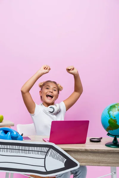 Escolar emocionado sentado cerca de la computadora portátil, globo y el arte de papel sobre fondo rosa - foto de stock