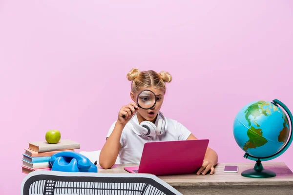 Écolier excité tenant loupe près d'un ordinateur portable, globe et papier d'art isolé sur rose — Photo de stock