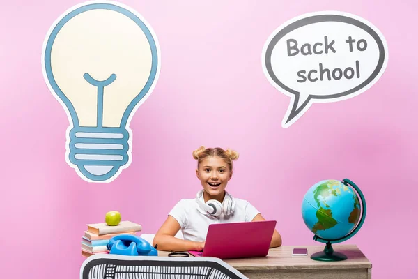 Child in headphones sitting near laptop, globe and paper artwork on pink background — Stock Photo