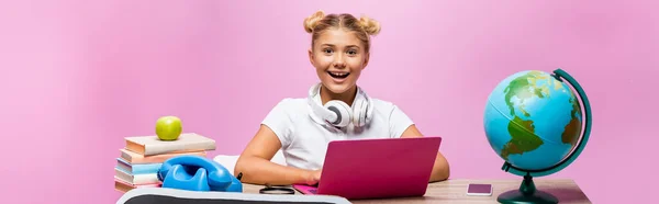 Cultura horizontal da criança em fones de ouvido sentado perto de gadgets, globo e livros sobre fundo rosa — Fotografia de Stock