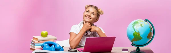 Colpo panoramico di scolara in cuffia seduta vicino a gadget, libri e globo su sfondo rosa — Foto stock