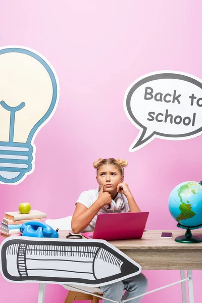 Pensive girl sitting near gadgets, globe with books and paper artwork on pink background — Stock Photo