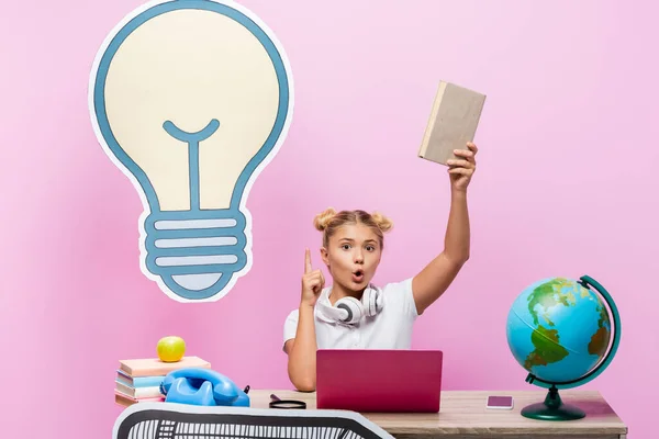 Excited kid having idea while holding book near gadgets and paper artwork on pink background — Stock Photo