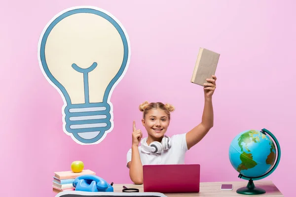 Kid pointing with finger while holding book near globe, gadgets and paper artwork on pink background — Stock Photo