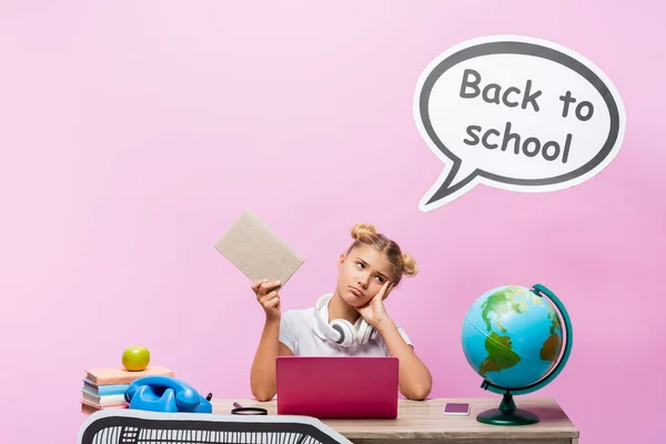 Tired kid holding book near gadgets, globe and speech bubble with back to school lettering on pink background — Stock Photo