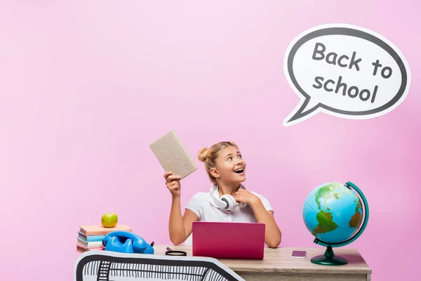 Colegiala sosteniendo libro y mirando la burbuja del habla con letras de vuelta a la escuela cerca de gadgets y globo sobre fondo rosa - foto de stock