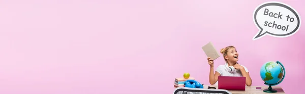Panoramic shot of kid holding book near globe, gadgets and paper artwork on pink background — Stock Photo