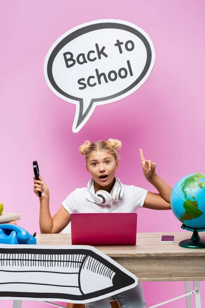 Schulmädchen mit Lupe, die auf Papierkunst in der Nähe von Gadgets, Globus und Büchern auf rosa Hintergrund zeigt — Stockfoto