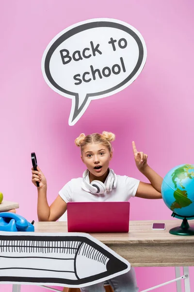 Colegiala con lupa apuntando al arte del papel cerca de gadgets, globo y libros sobre fondo rosa - foto de stock