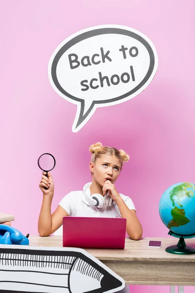 Colegiala pensativa con lupa cerca de gadgets, libros y arte de papel sobre fondo rosa - foto de stock