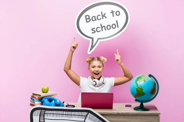 Studentessa indicando bolla discorso con di nuovo a scuola lettering vicino globo, gadget e libri su sfondo rosa — Foto stock