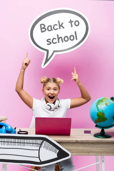 Muchacho emocionado apuntando a la burbuja del habla con letras de vuelta a la escuela cerca de la computadora portátil, teléfono inteligente y globo en el escritorio sobre fondo rosa - foto de stock