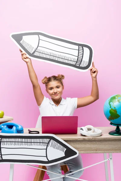 Schoolgirl holding paper pencil near laptop, globe and telephone on pink background — Stock Photo
