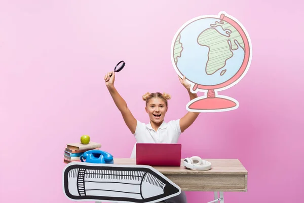 Excited schoolkid holding loupe and paper globe near laptop, headphones and books on pink background — Stock Photo