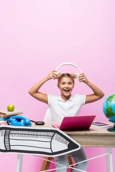 Écolière excitée tenant des écouteurs près de gadgets, livres et illustrations en papier sur fond rose — Photo de stock