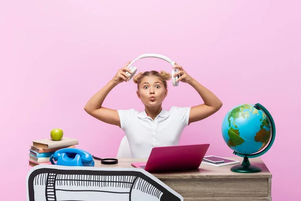 Écolière soufflant l'air baiser tout en tenant écouteurs près de gadgets, livres et illustrations en papier sur fond rose — Photo de stock