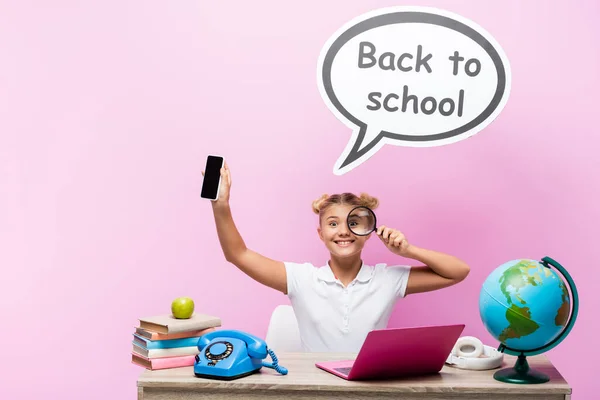 Schoolkid holding magnifying glass and smartphone near books, laptop and speech bubble with back to school lettering on pink background — Stock Photo