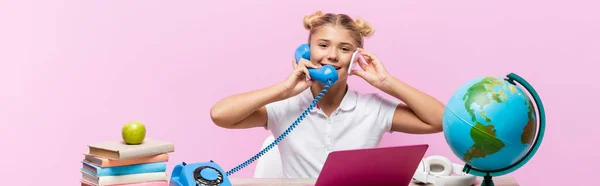 Panoramic shot of schoolgirl talking on telephone and smartphone near laptop and globe isolated on pink — Stock Photo