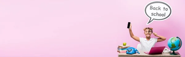 Colpo panoramico di scolaro che tiene smartphone e lente d'ingrandimento vicino al globo e bolla vocale con iscrizione a scuola su sfondo rosa — Foto stock