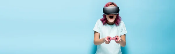 QUIIV, UCRÂNIA - JULHO 29, 2020: jovem excitada com cabelo rosa no fone de ouvido vr jogando videogame com joystick em fundo azul, tiro panorâmico — Fotografia de Stock