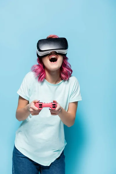 KYIV, UKRAINE - 29 juillet 2020 : jeune femme excitée avec des cheveux roses dans un casque vr jouer à un jeu vidéo avec joystick sur fond bleu — Photo de stock