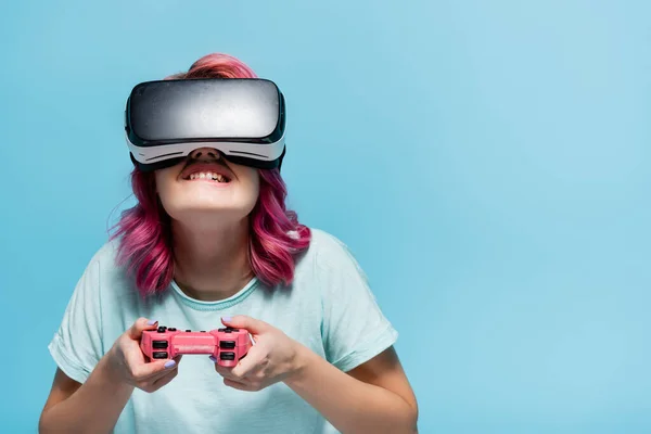 KYIV, UKRAINE - JULY 29, 2020: concentrated young woman with pink hair in vr headset playing video game with joystick isolated on blue — Stock Photo