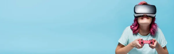 KIEW, UKRAINE - 29. JULI 2020: Konzentrierte junge Frau mit pinkfarbenen Haaren im vr-Headset, die Videospiel mit Joystick spielt, isoliert auf blauem Hintergrund, Panoramaaufnahme — Stockfoto