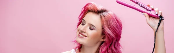 Young woman with colorful hair and closed eyes holding straightener isolated on pink, panoramic shot — Stock Photo