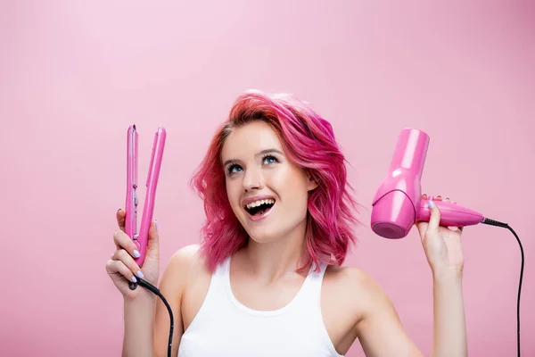 Aufgeregte junge Frau mit buntem Haar, Glätteisen und Haartrockner isoliert auf rosa — Stockfoto