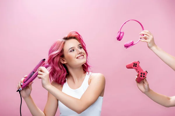 KYIV, UKRAINE - JULHO 29, 2020: jovem com cabelos coloridos usando alisador perto de fones de ouvido e joystick em mãos isoladas em rosa — Fotografia de Stock