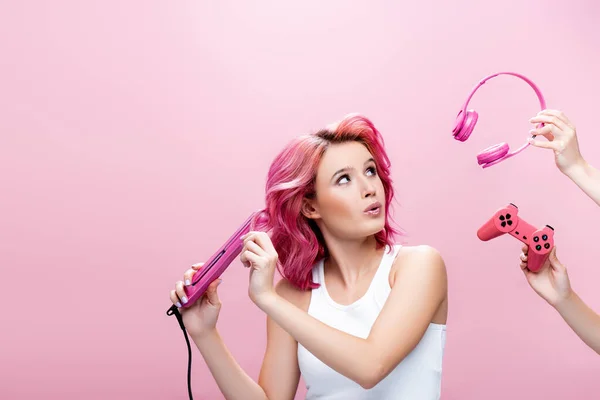KYIV, UCRAINA - 29 LUGLIO 2020: giovane donna sorpresa con capelli colorati utilizzando piastra vicino alle cuffie e joystick in mani isolate su rosa — Foto stock